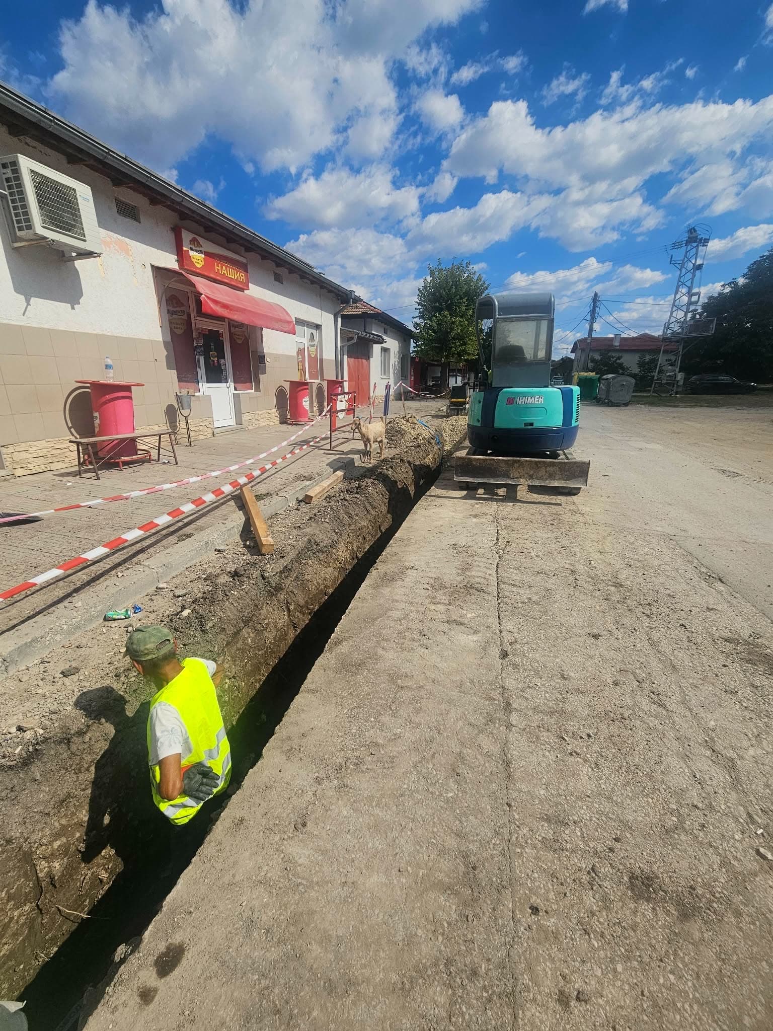Дълъг изкоп покрай тротоара в центъра на село, обезопасен с лента; работник в изкопа и мини багер на улицата.